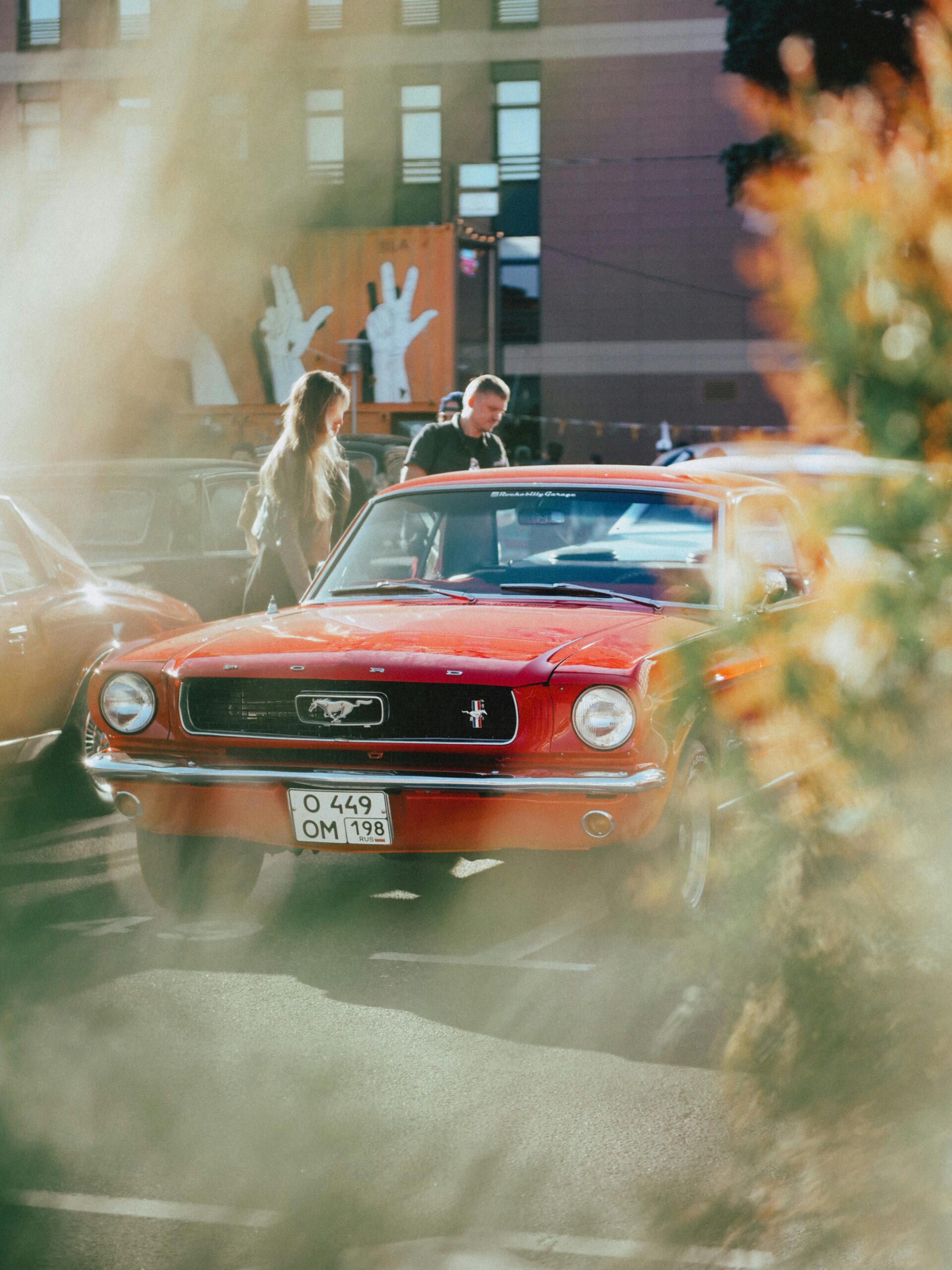 A classic red Ford Mustang parked outdoors with people nearby in a city setting.